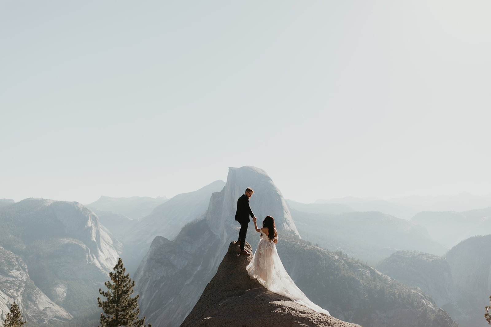 Groom helping his bride up a granite peak with Half Dome in the background, an adventurous mountain elopement moment.
