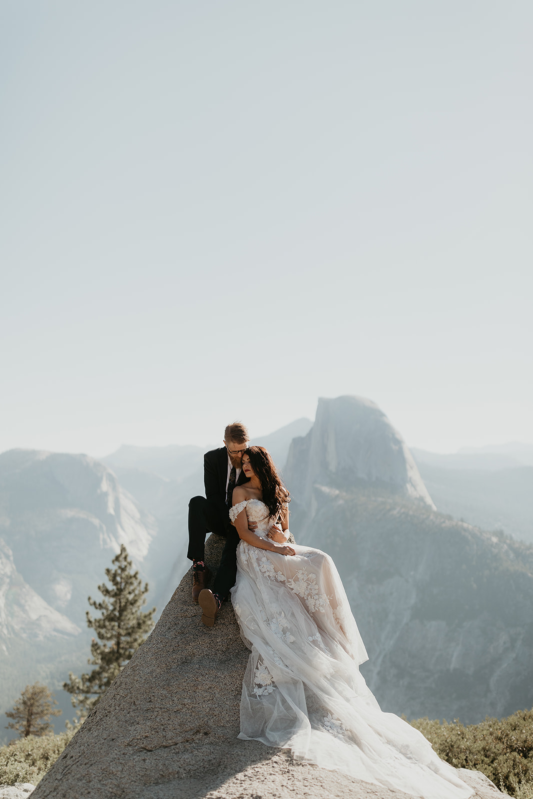 Bride and groom sitting atop a rock with panoramic views of Half Dome in the distance, elopement-style elegance.