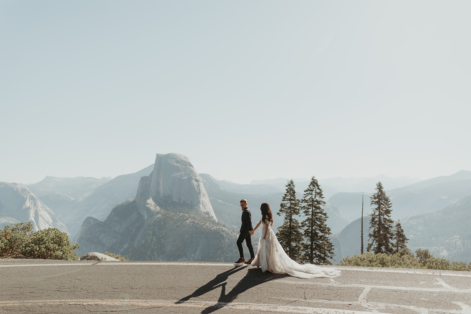 Bride and groom walking along a road with sweeping views of Yosemite and Half Dome behind them.