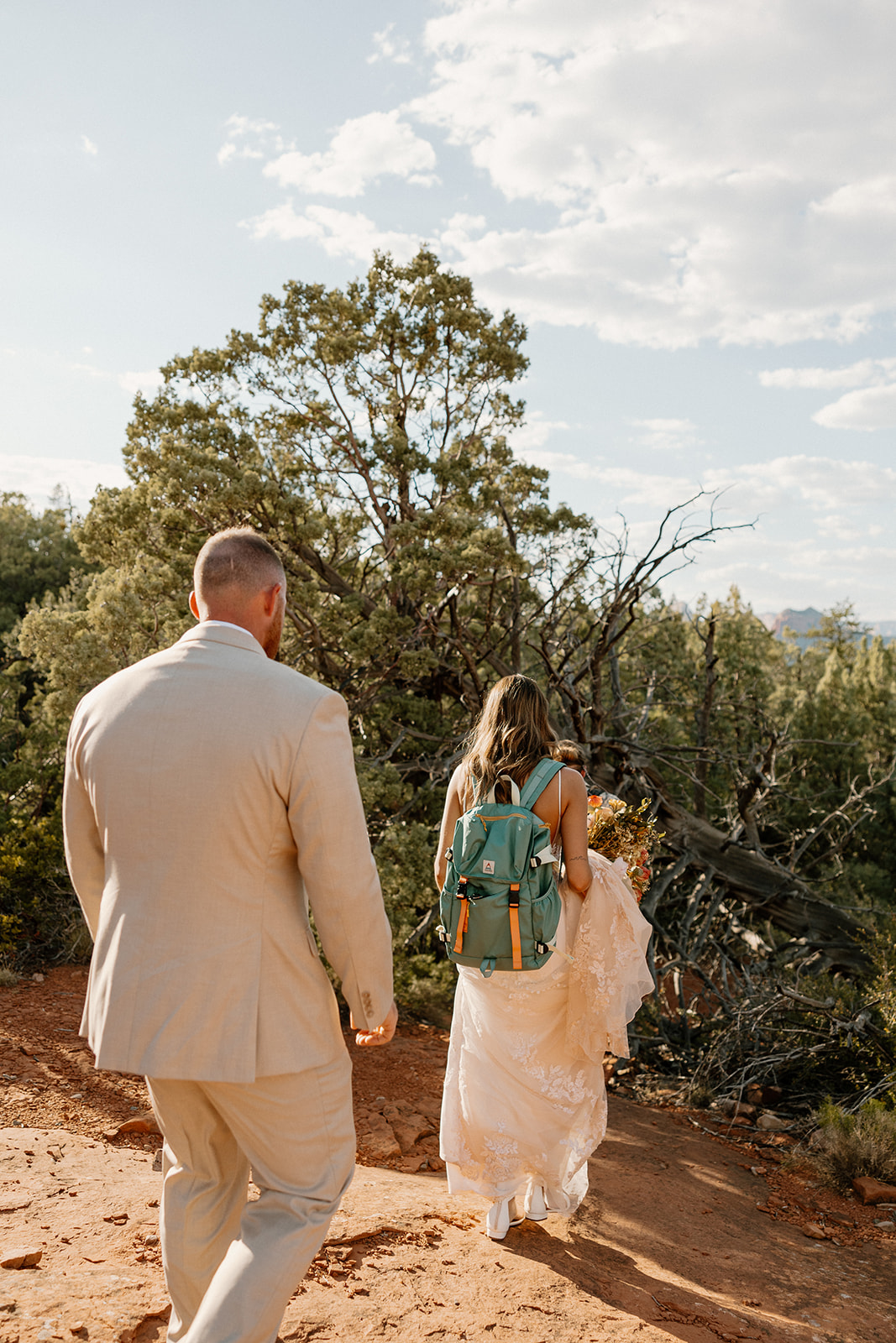 Bride with a green hiking backpack and groom walking a red dirt trail, heading to their Arizona elopement ceremony site.