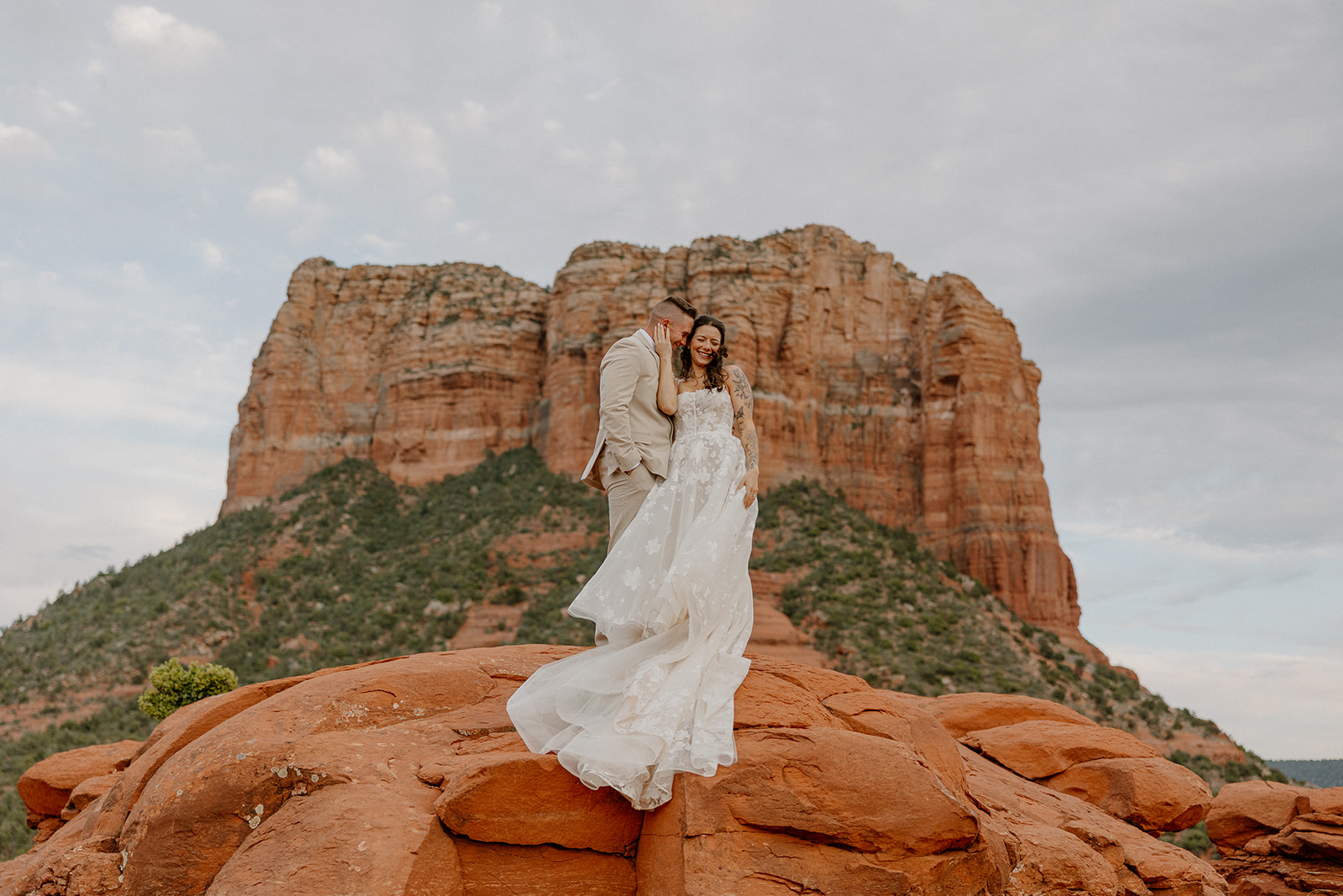 Bride in flowing floral wedding attire laughing with groom in front of towering red rock backdrop

