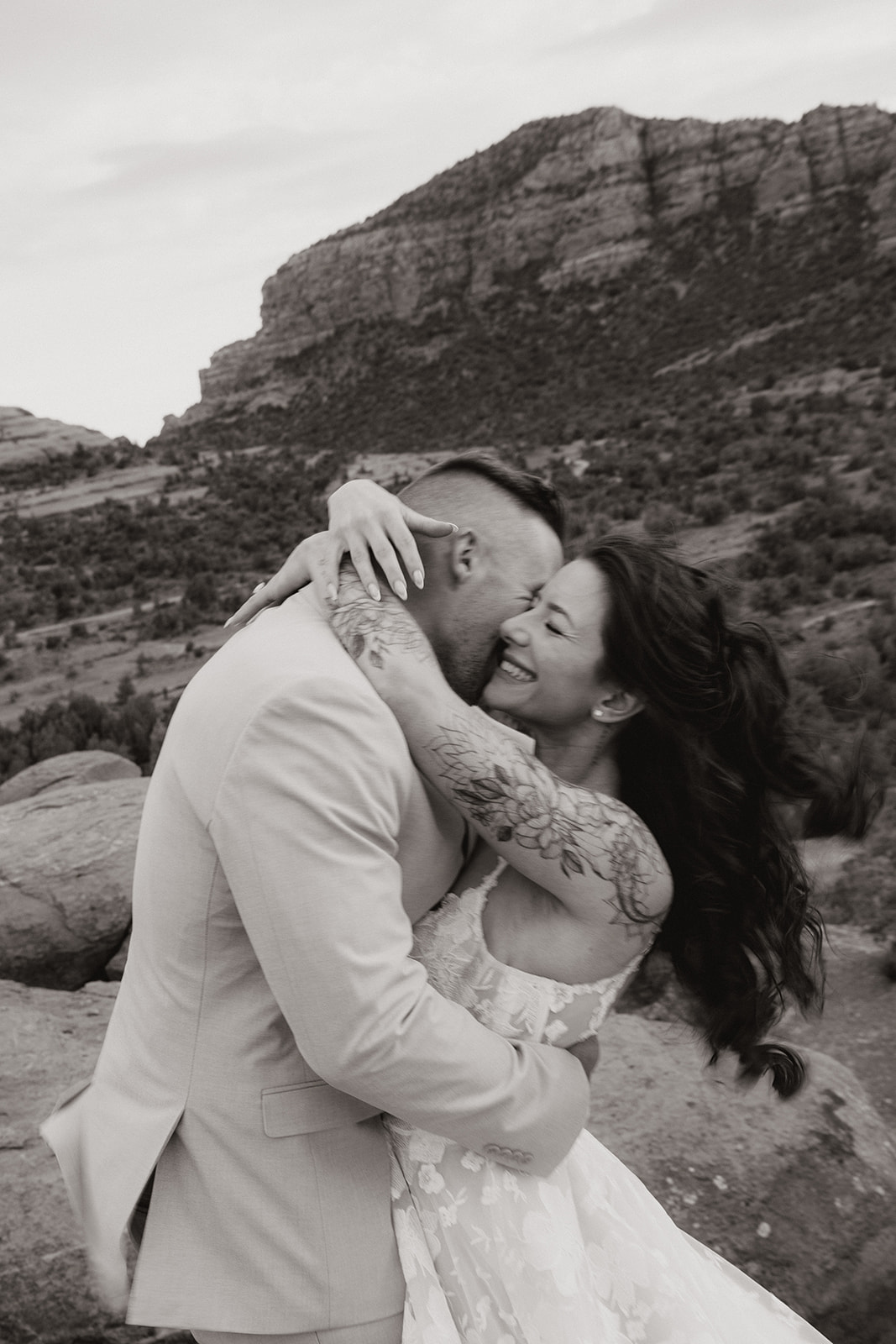 Bride and groom locked in a joyful embrace with mountains behind them, captured in candid black and white.