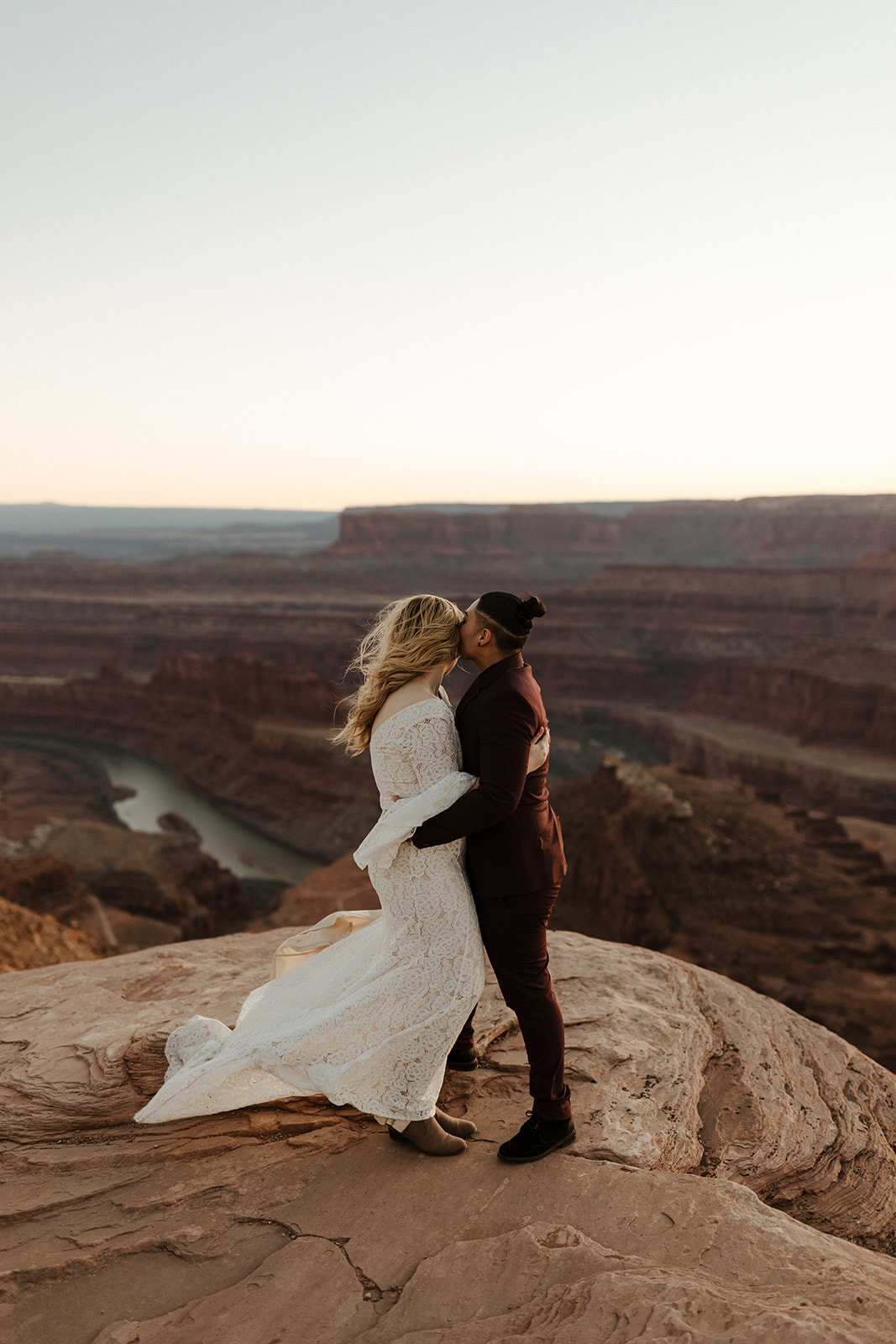 Couple sharing a kiss on a windswept cliff above a winding river at sunset, wrapped in soft desert light.