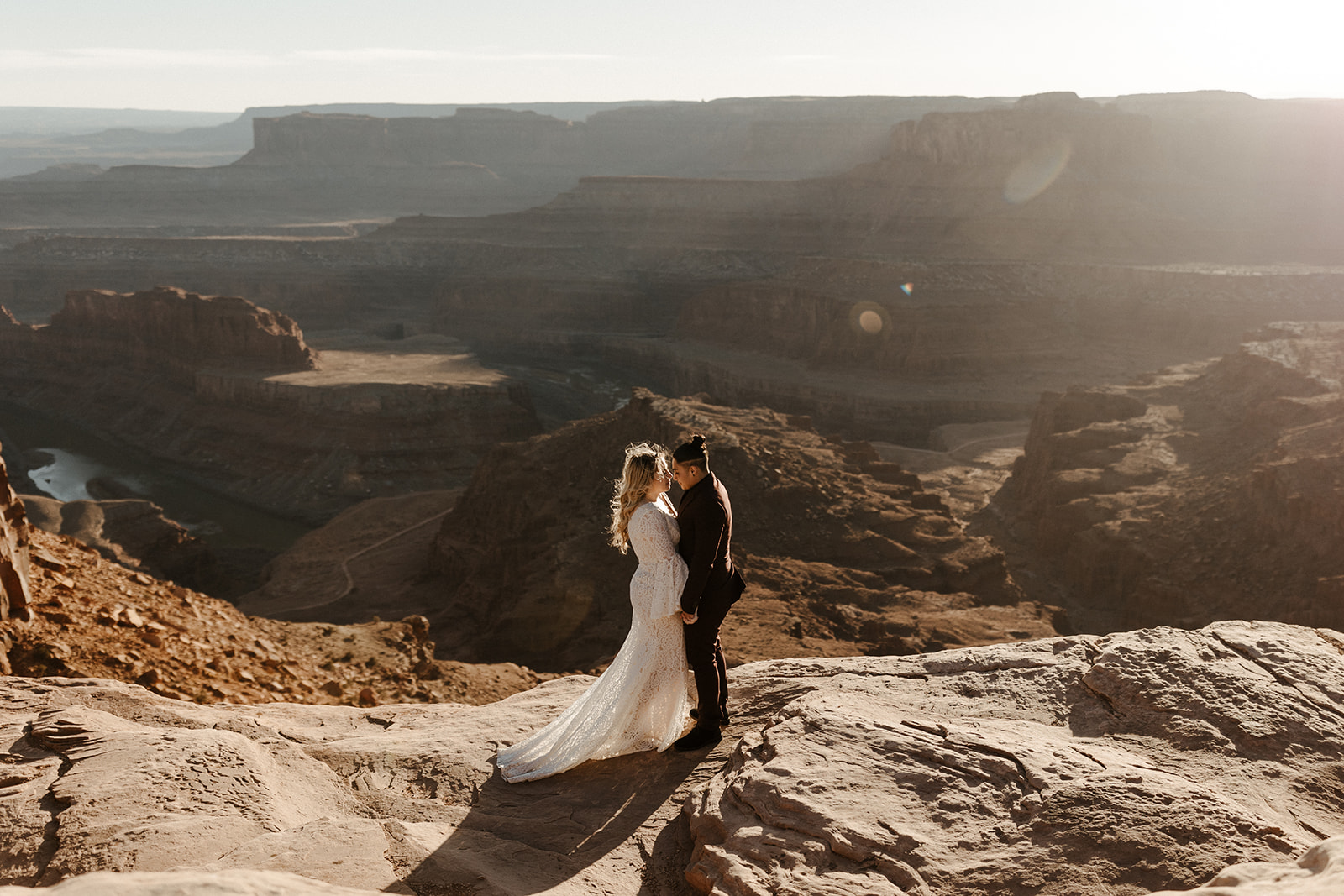 Couple holding hands and gazing at each other on a canyon cliff at sunset, desert elopement vibes.
