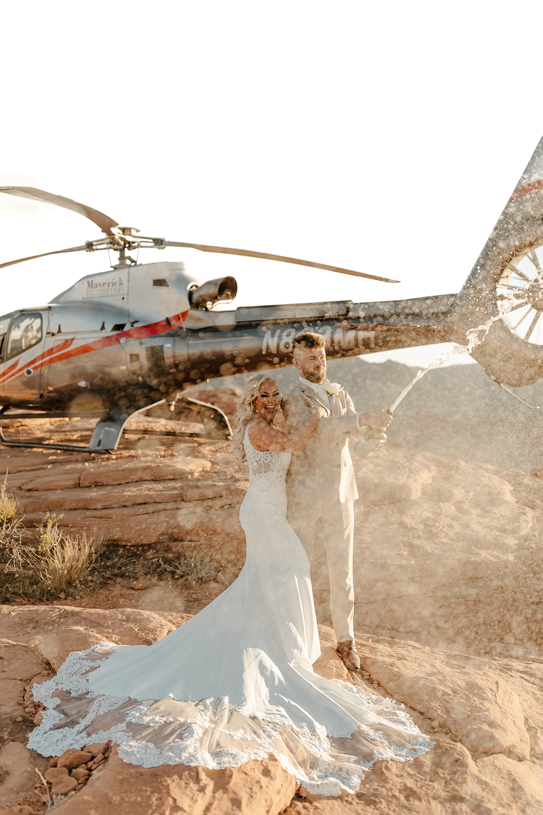 Bride and groom in sleek wedding attire popping champagne in front of a helicopter on red rock terrain
