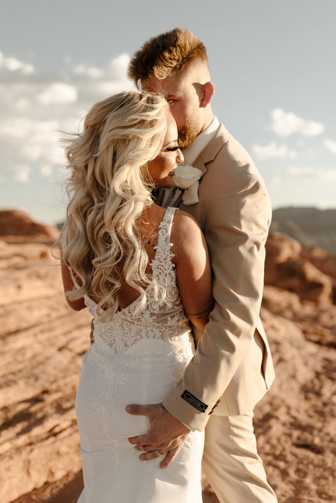 Groom in tan suit embracing bride in intricate lace wedding attire on sunlit red rocks

