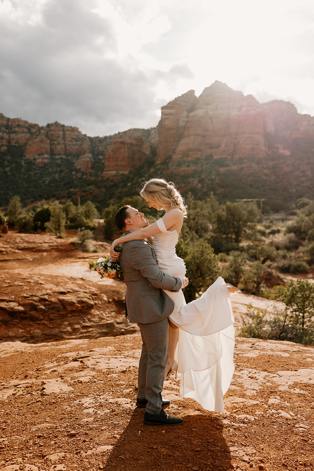 Groom lifting bride in front of dramatic red rock formations in Sedona during their joyful Arizona elopement.
