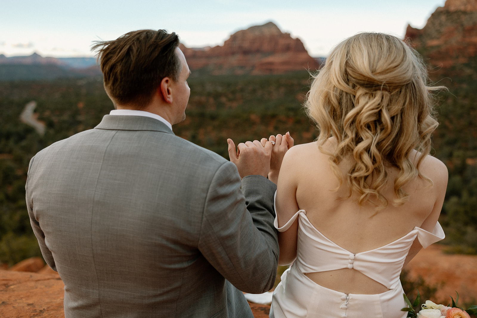 Bride and groom linking pinkies, seated on red rock with Sedona’s iconic formations in the distance.
