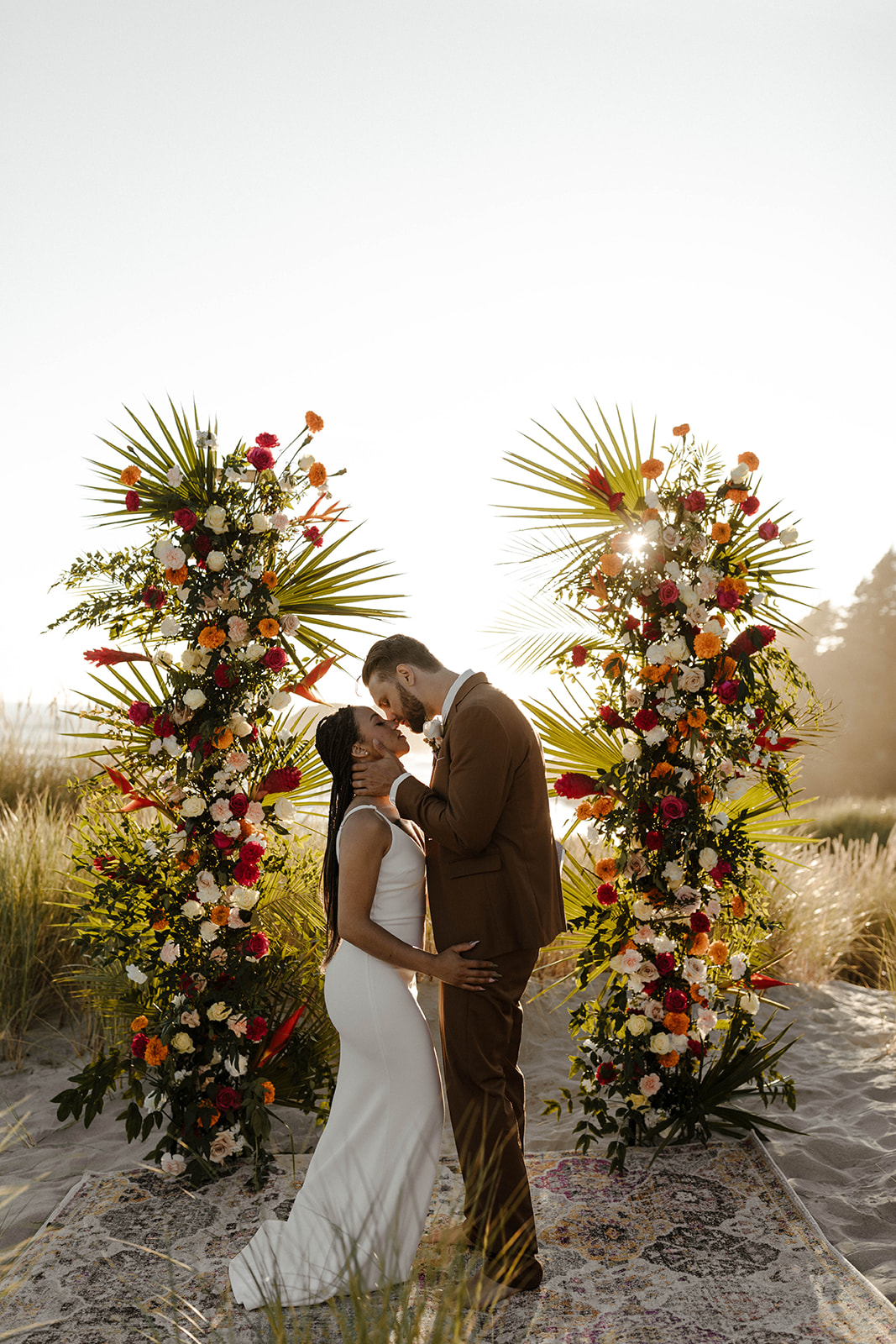 Colorful floral pillars frame newlyweds kissing on a vintage rug at their sunset beach wedding