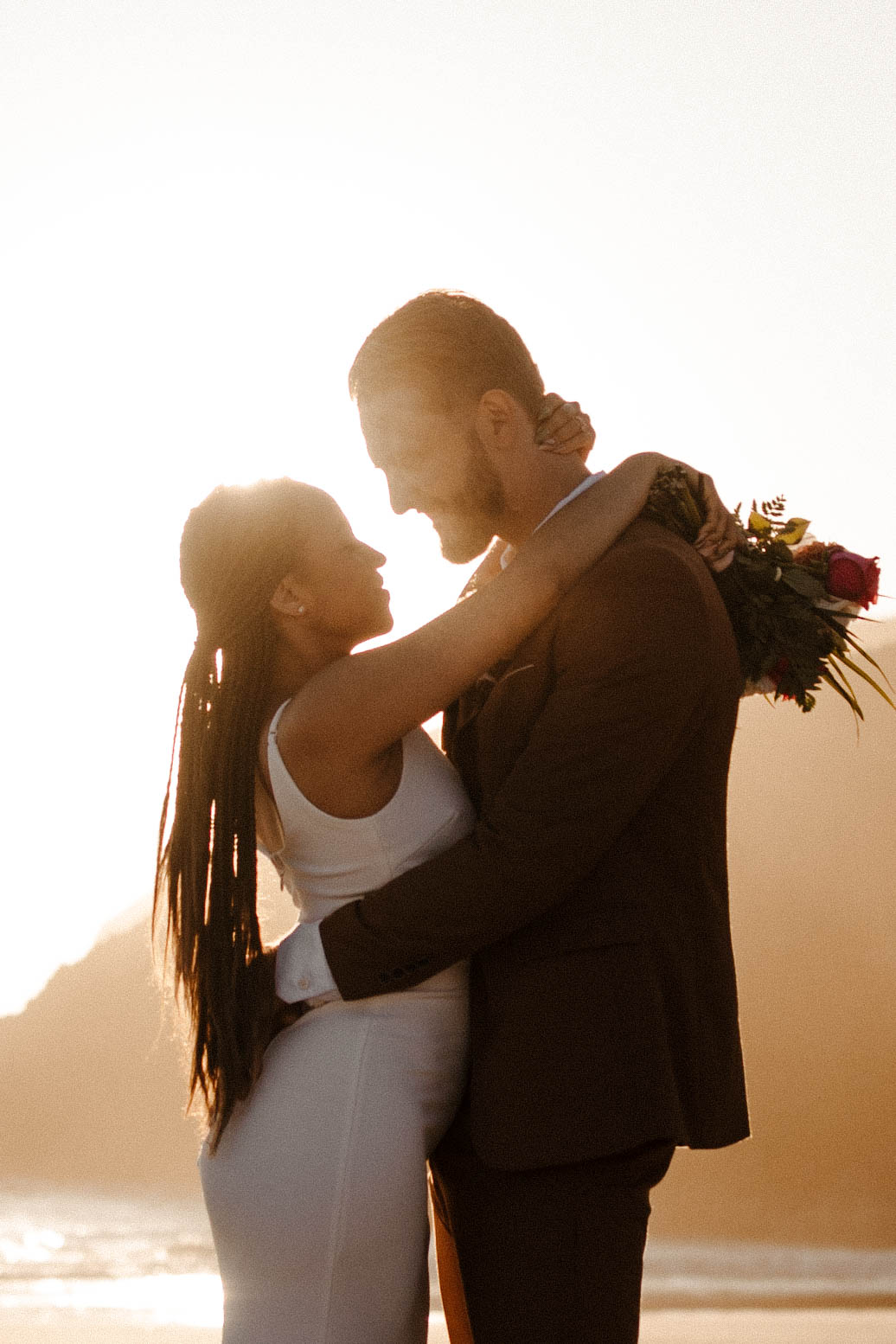Silhouetted couple holding each other at sunset on the beach, radiating warmth and peace after their private elopement.
