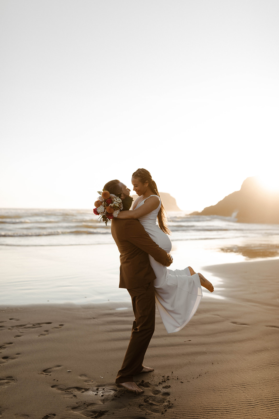 Groom lifting barefoot bride in sleek modern wedding attire as waves crash behind them