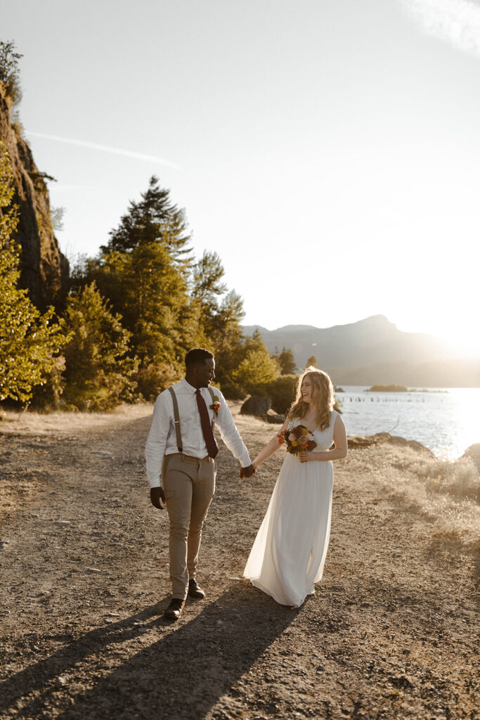 Newlyweds walking hand in hand on a gravel trail beside a lake, glowing with joy after their low-key wedding ceremony.
