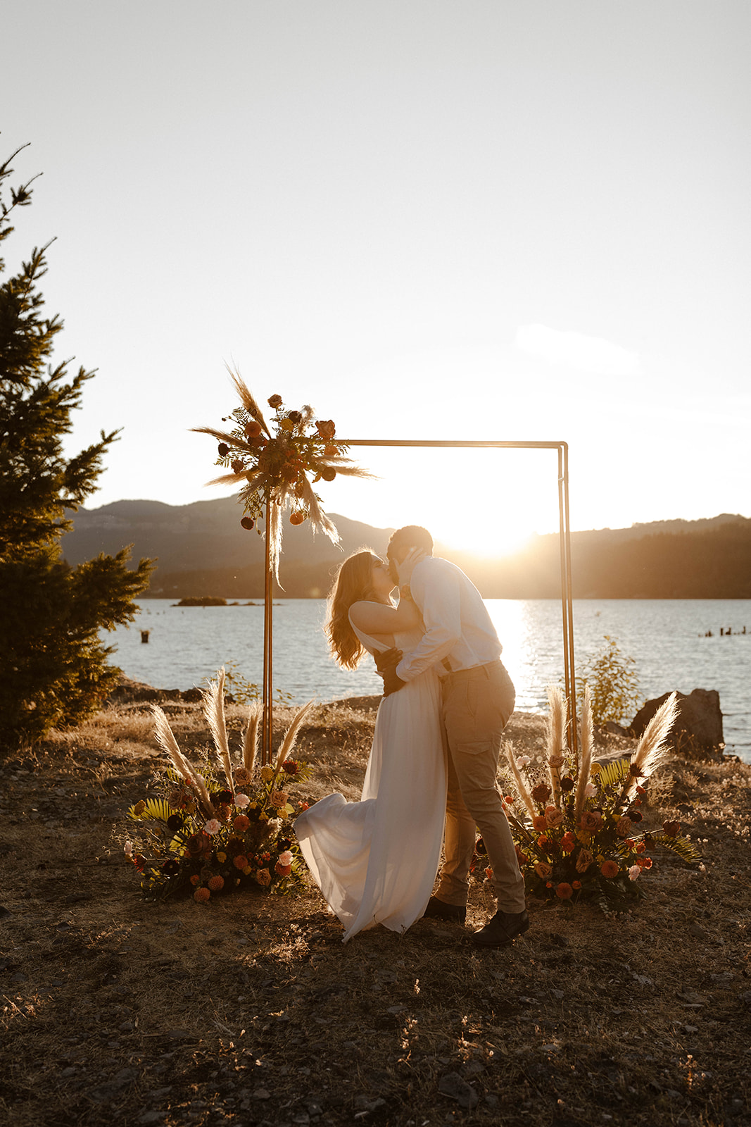 A romantic sunset kiss beneath a floral arch by the water, proving what is the best time of day to elope when you want glowy, golden vibes.