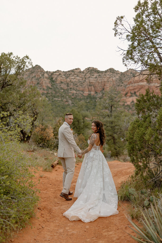 Bride in embroidered wedding attire looking over her shoulder as she and her groom walk a red dirt path in Sedona

