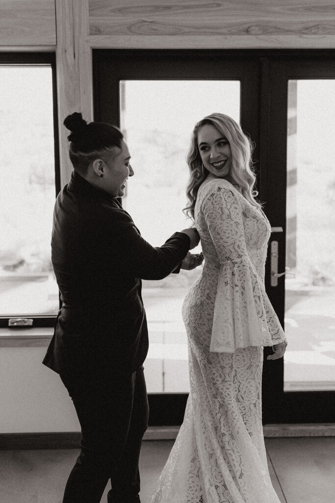 Black and white image of bride in lace gown smiling as partner adjusts her sleeve indoors