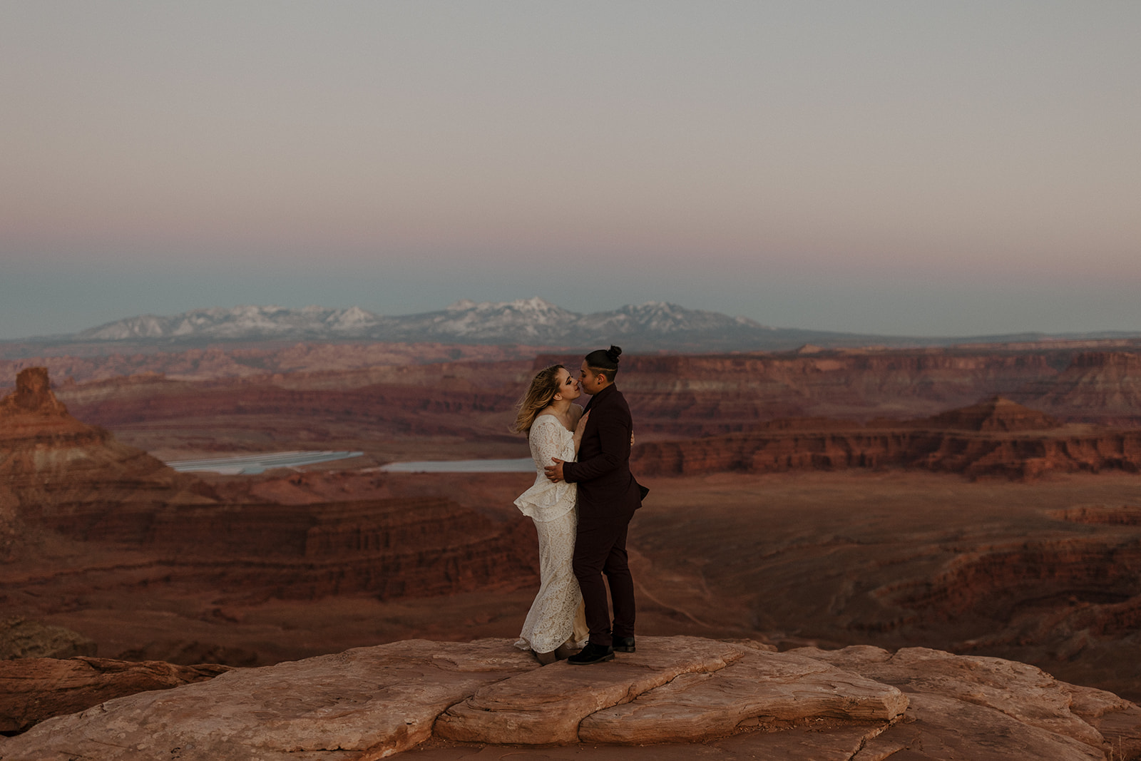 Groom wedding attire featuring a dark maroon suit styled for a cliffside elopement at Dead Horse Point, with soft pastels in the sunset sky.