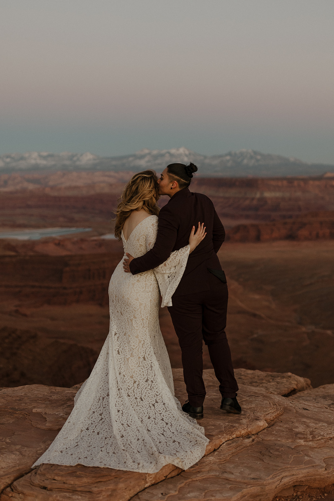 Two brides embracing at sunset on a canyon cliff, golden hour glow highlighting their elopement celebration.
