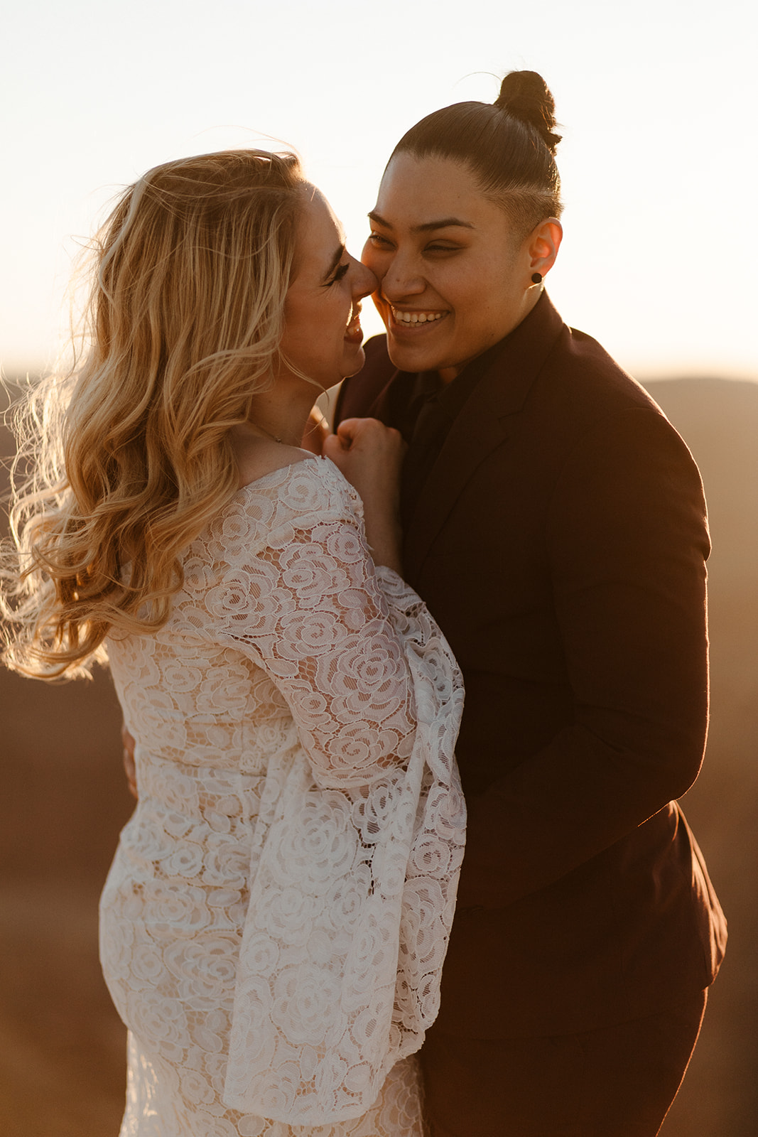 Golden hour laughter between newlyweds, one in a lace dress, the other in a burgundy suit

