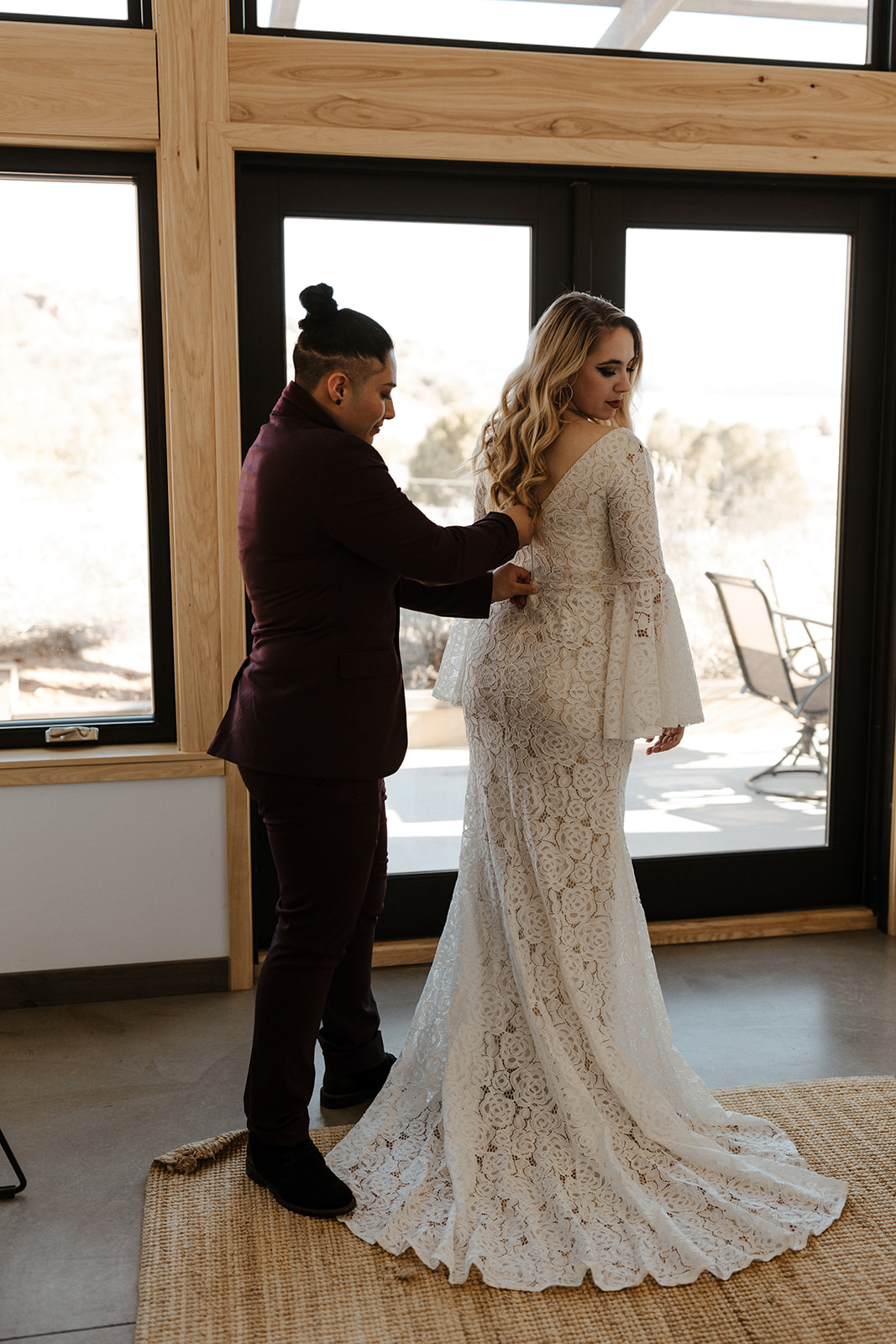 Getting ready moment with one partner helping the other into a bell-sleeve lace dress, a beautiful pause before telling family they're eloping.
