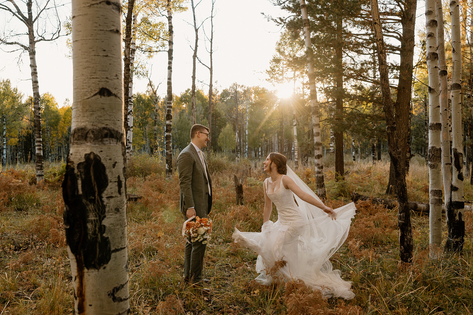 Bride playfully lifting her dress and laughing with her groom in a sun-dappled forest, filled with post-ceremony joy.
