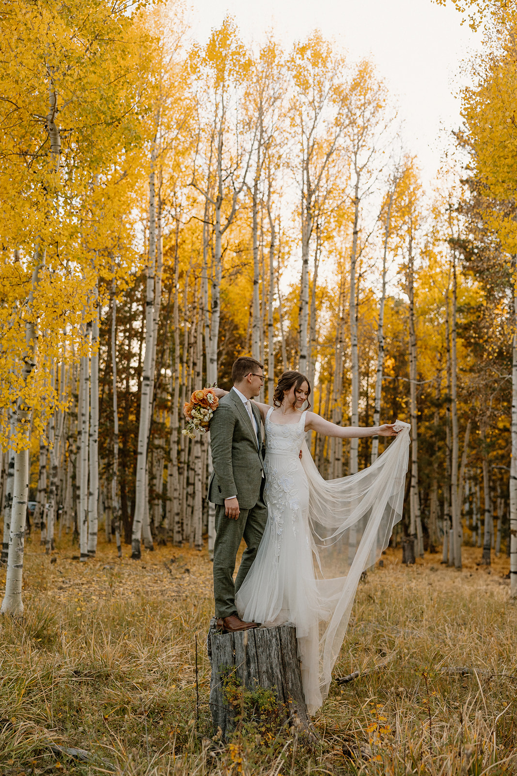 Autumn elopement moment in a golden aspen grove, bride lifting her dress while standing beside her groom on a tree stump.
