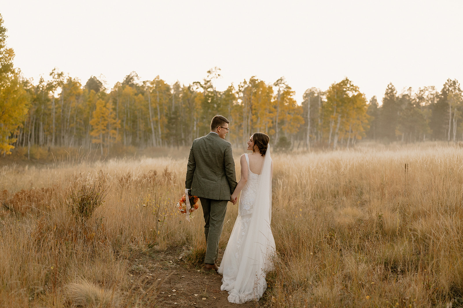 Bride and groom holding hands and walking through a golden meadow, with fall trees in the background and just-married smiles on their faces.
