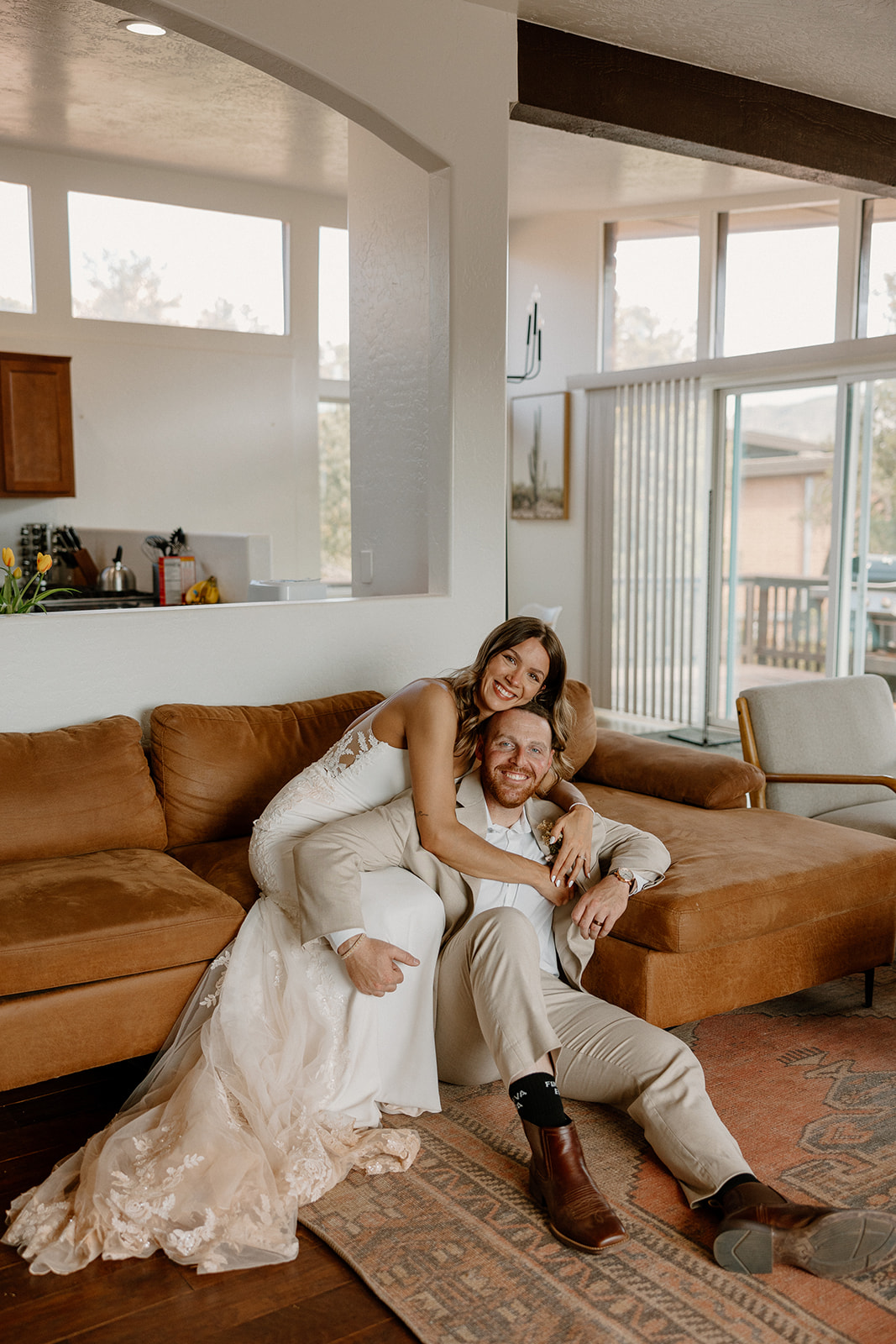 Bride in a lace gown wrapping her arms around her partner on a cozy brown sectional, capturing a laid-back moment after navigating how to tell your family you're eloping.
