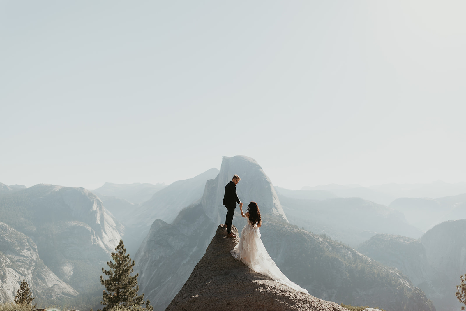 Epic elopement shot of couple on a rock ledge in Yosemite, holding hands with Half Dome in the distance.
