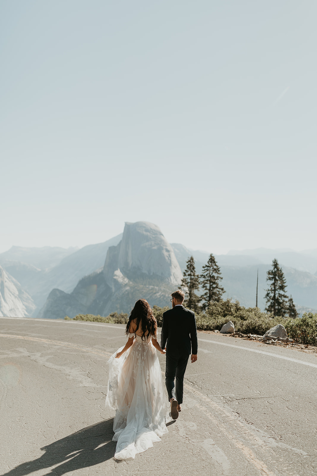 Bride and groom walking down a quiet mountain road in Yosemite, hand in hand with Half Dome in the distance—adventuring into married life their way.
