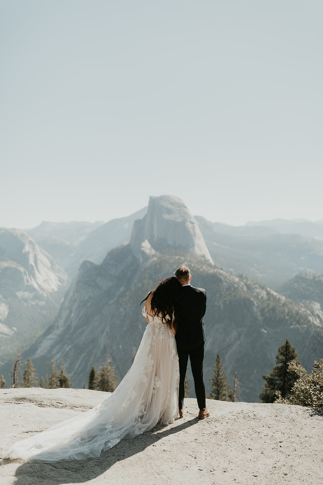 Back view of groom wedding attire in a tailored black suit, holding his bride close as they overlook Half Dome from a Yosemite vista.
