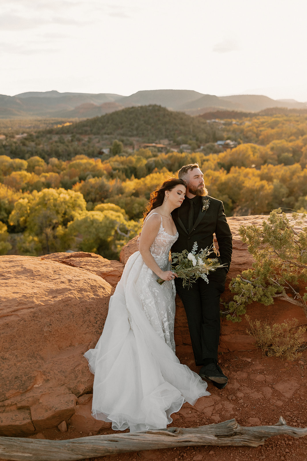 Bride and groom sitting on red rock with arm around one another watching the sunset
