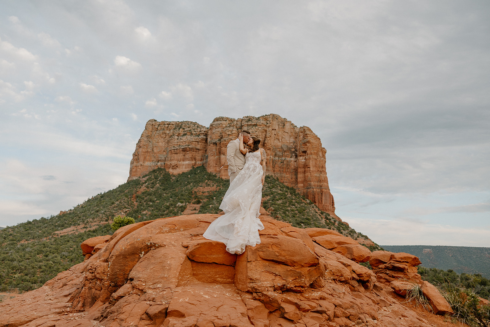 Bride touching groom's cheek while wind blows through her dress and hair while standing on red rocks in Arizona