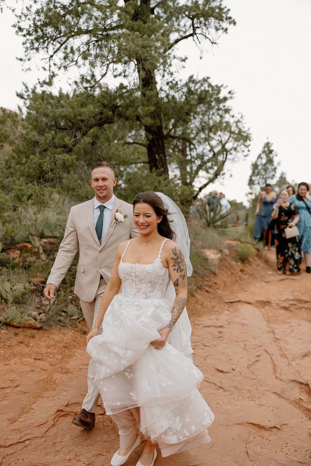 Bride lifting her dress with a big smile, walking the red dirt path with her groom and guests after their Arizona elopement.