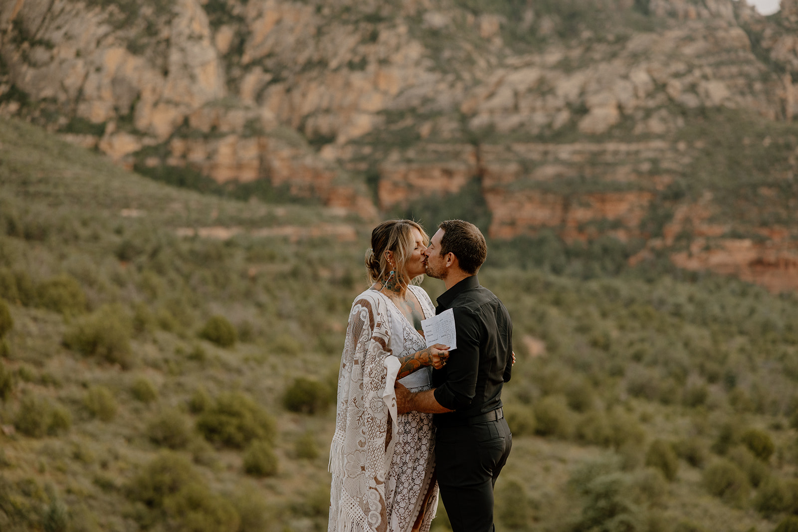 Bride in fringe-trimmed lace wedding attire kisses her groom after reading vows in the red rock canyon

