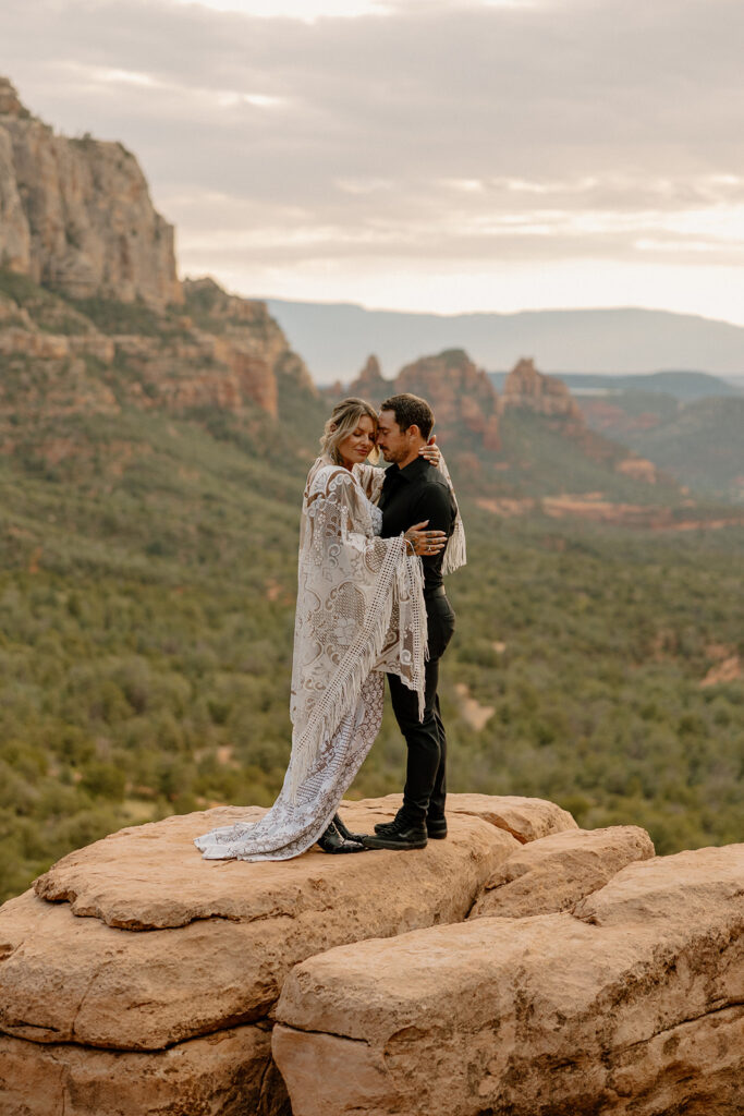 Couple standing arm-in-arm on a rock ledge, bride’s boho wedding attire catching the breeze

