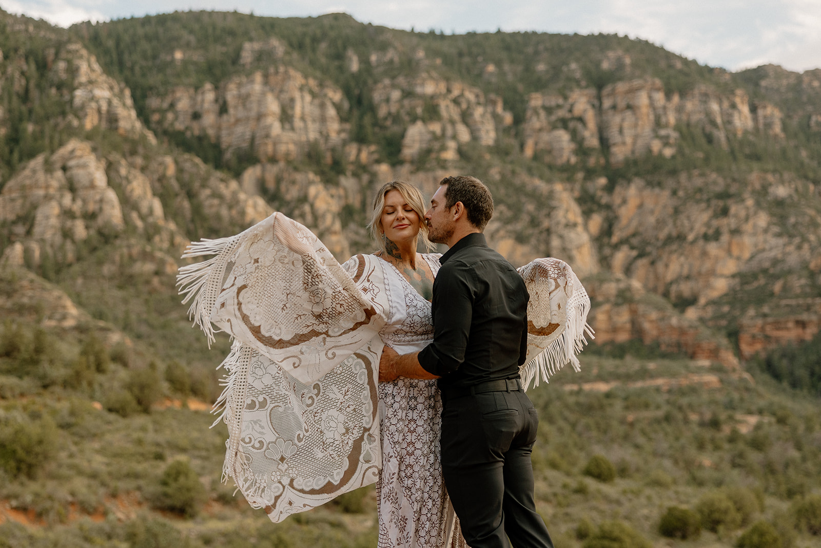 Bride in fringed lace wedding attire flowing in the breeze as groom kisses her cheek against canyon cliffs