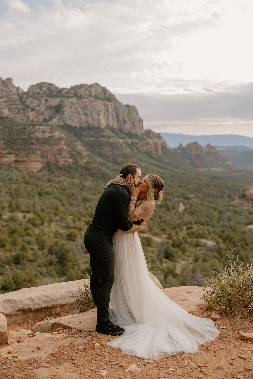 Bride in airy tulle wedding attire shares a joyful kiss with her partner atop a Sedona overlook

