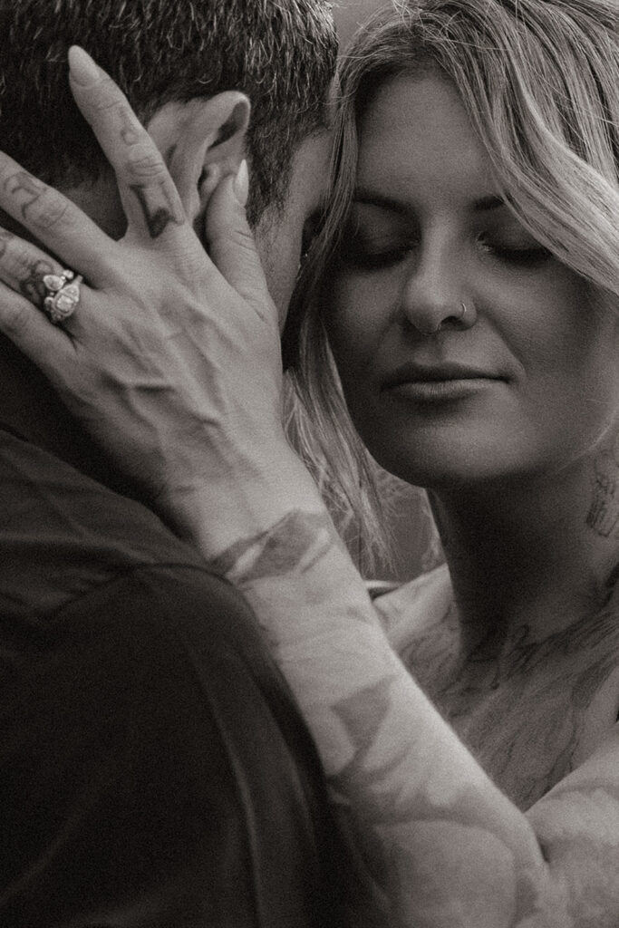 Black and white close-up of bride gently cradling her partner’s face, showcasing her ring and tattoos