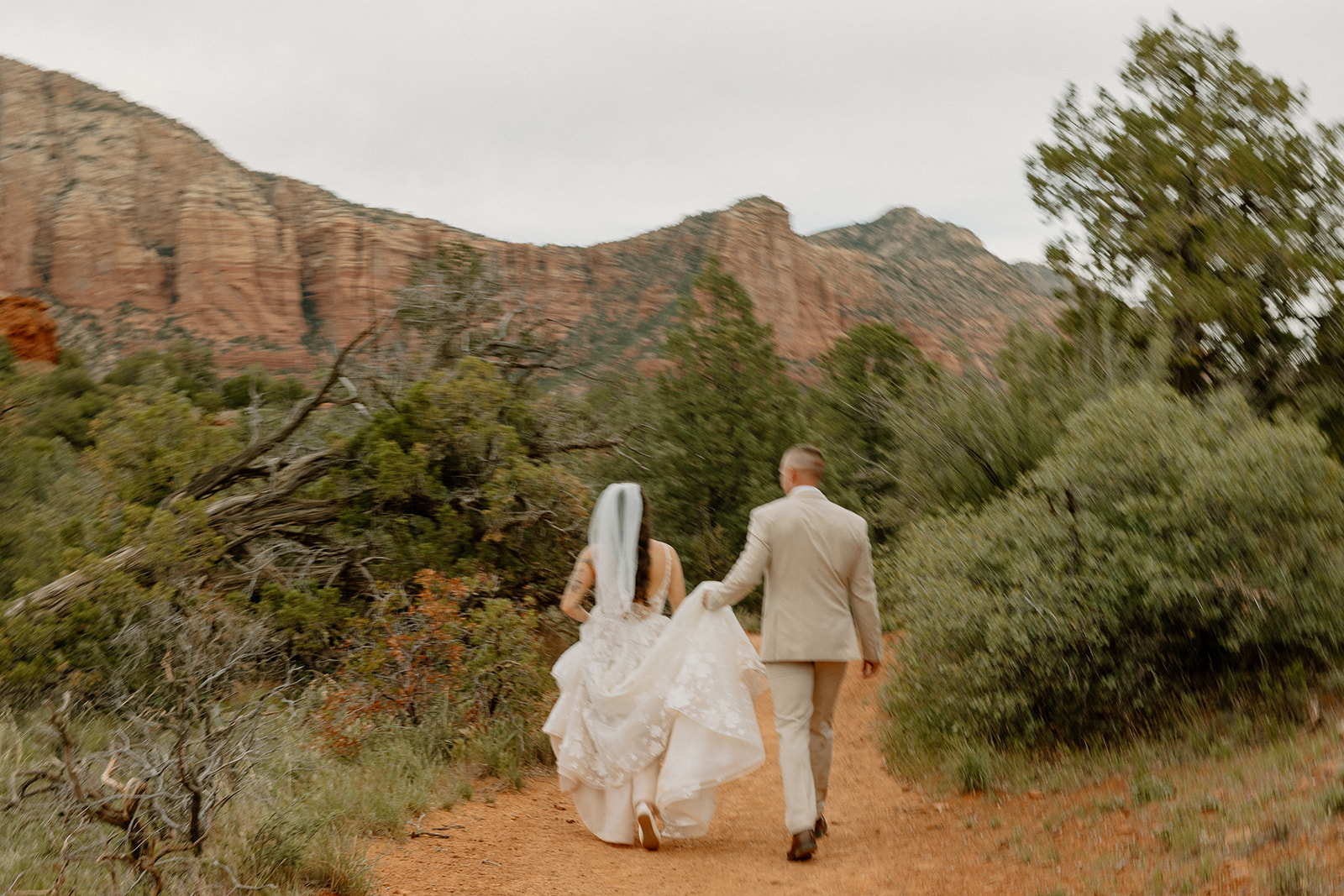 Couple walking hand in hand down a Sedona trail, bride lifting her gown to keep it clean