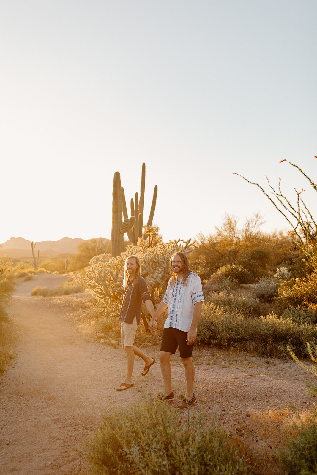 wo grooms walk hand-in-hand on a glowing desert trail during golden hour, showcasing what is the best time of day to elope for dreamy desert light.