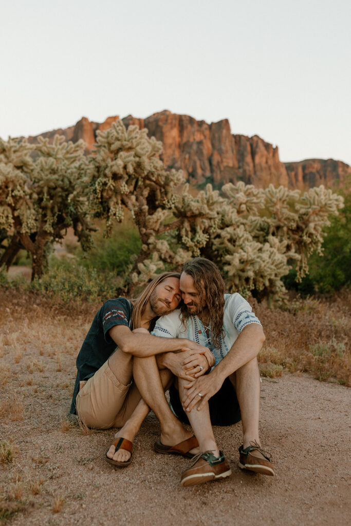 Two grooms sitting close together under cholla cacti, laughing during their desert elopement