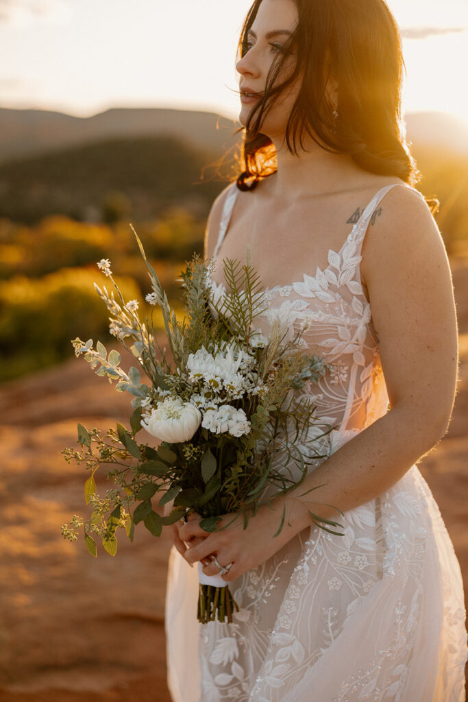 Close-up of bride holding wildflower bouquet in sheer embroidered gown, glowing in sunset light

