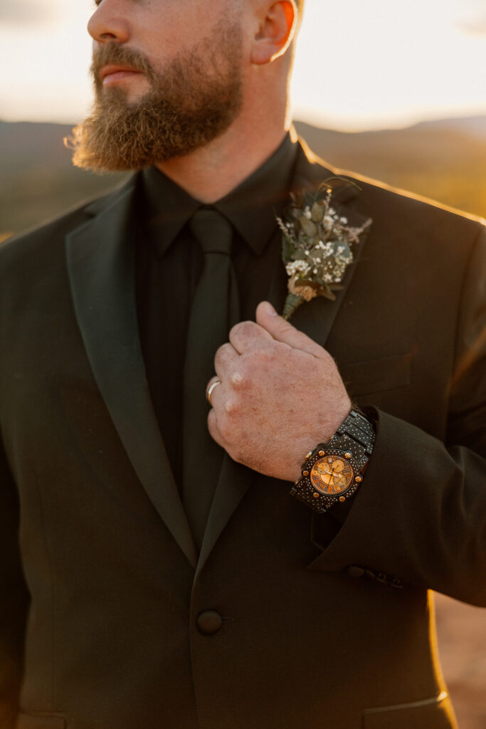 Groom in black suit and tie with gold watch and boutonnière, highlighting modern wedding attire details