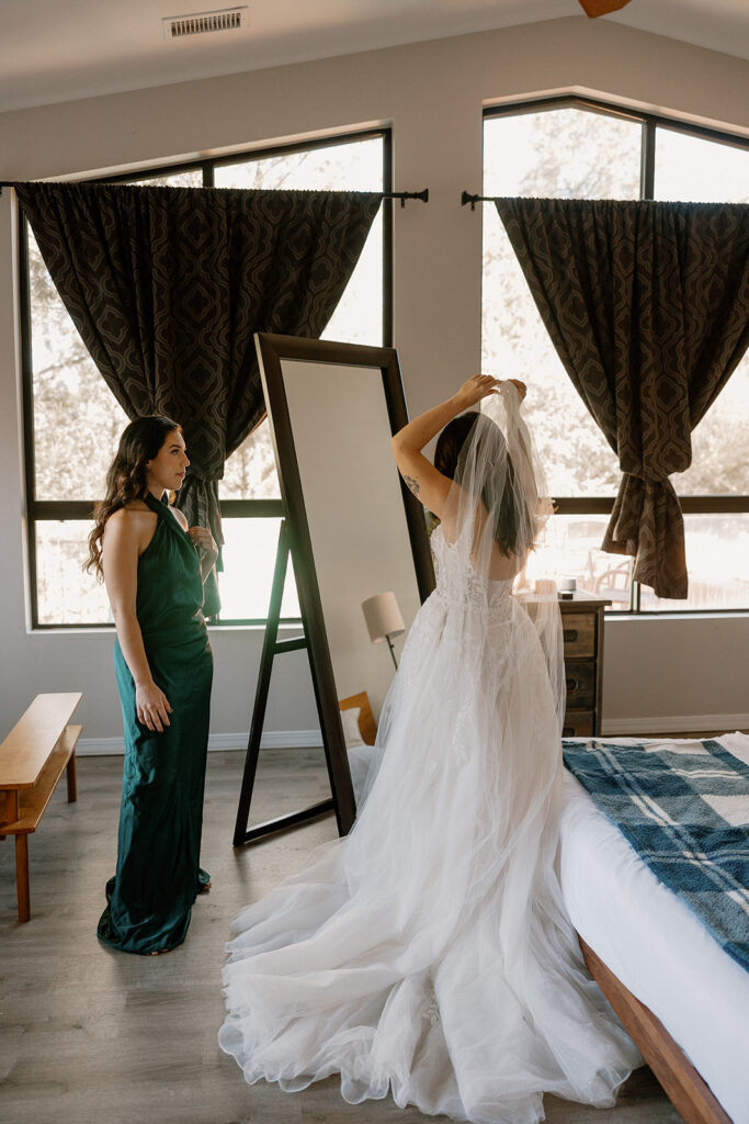 Bride adjusting her veil in front of a full-length mirror while bridesmaid watches in natural light

