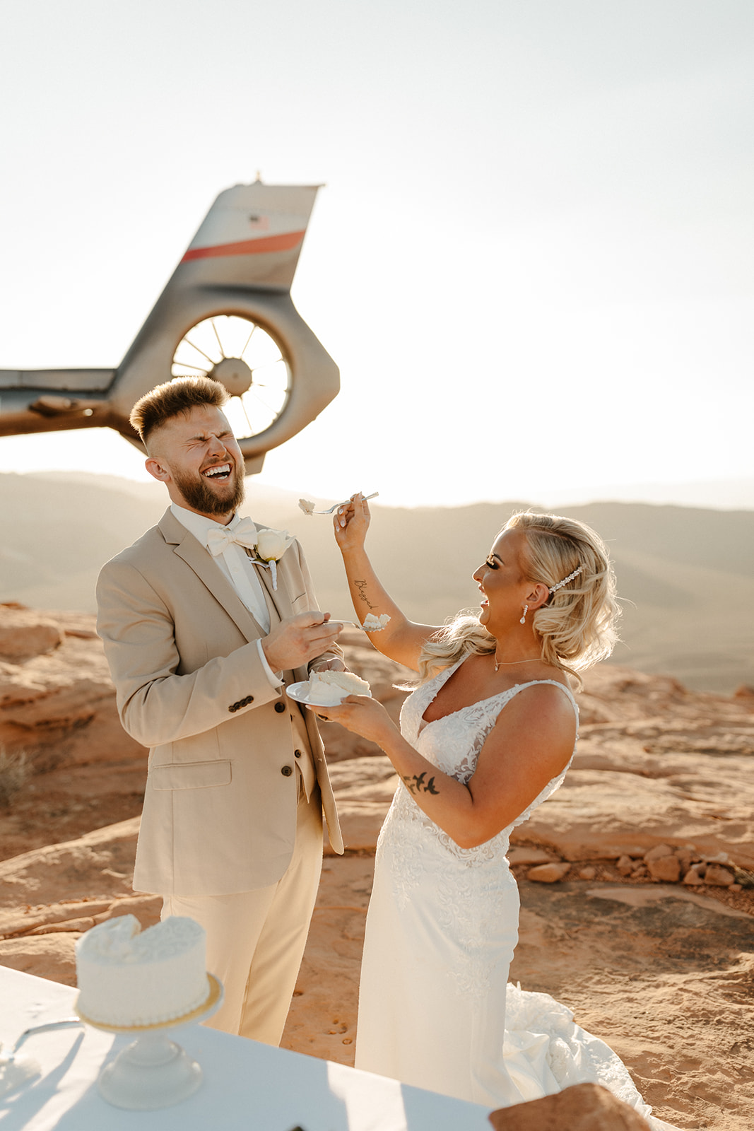 Adventurous cake-cutting celebration with groom wedding attire in a beige suit, sharing a laugh with his bride near a helicopter landing.
