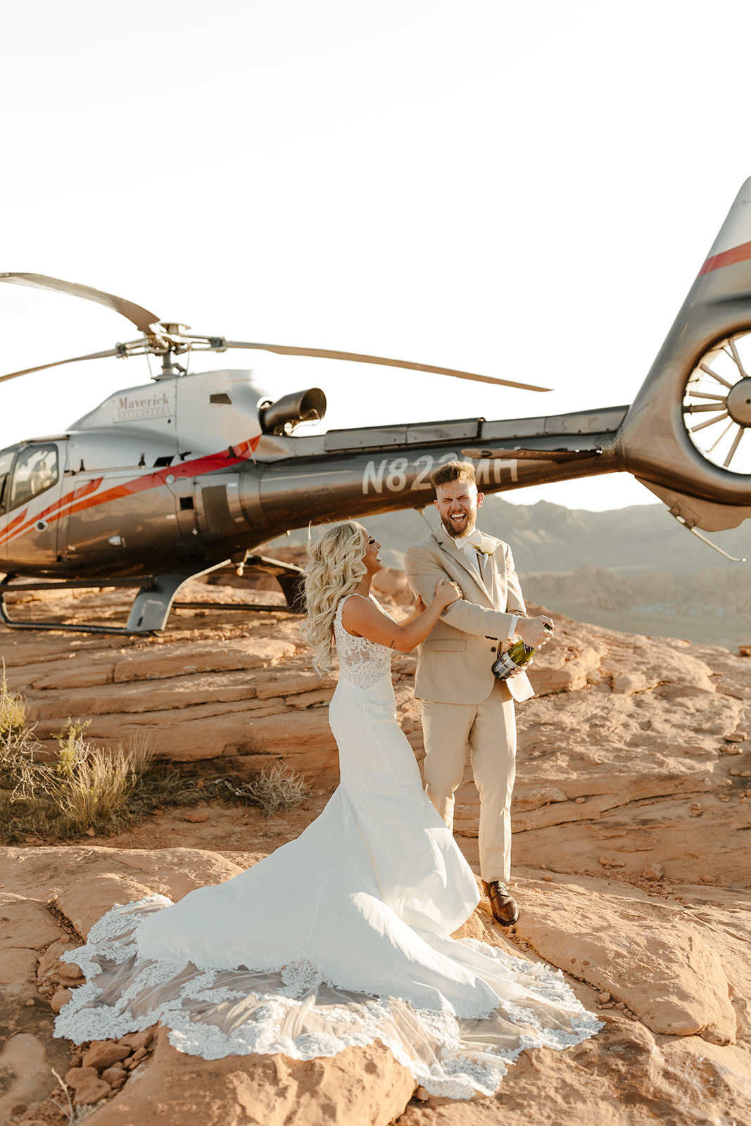 Desert elopement celebration with groom wedding attire in a beige suit and leather shoes, popping champagne beside a helicopter on the red rocks.