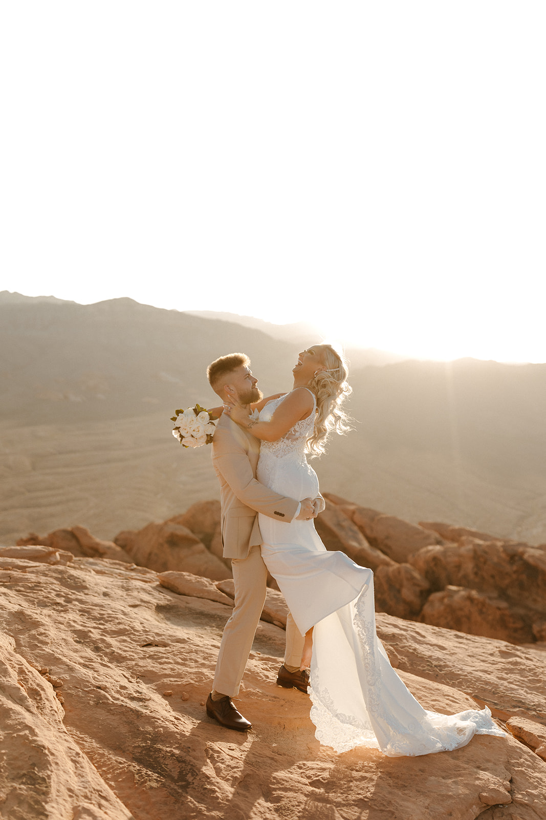 Sun-drenched cliffside elopement with groom wedding attire in a beige suit, lifting the bride for a joyful sunset kiss.