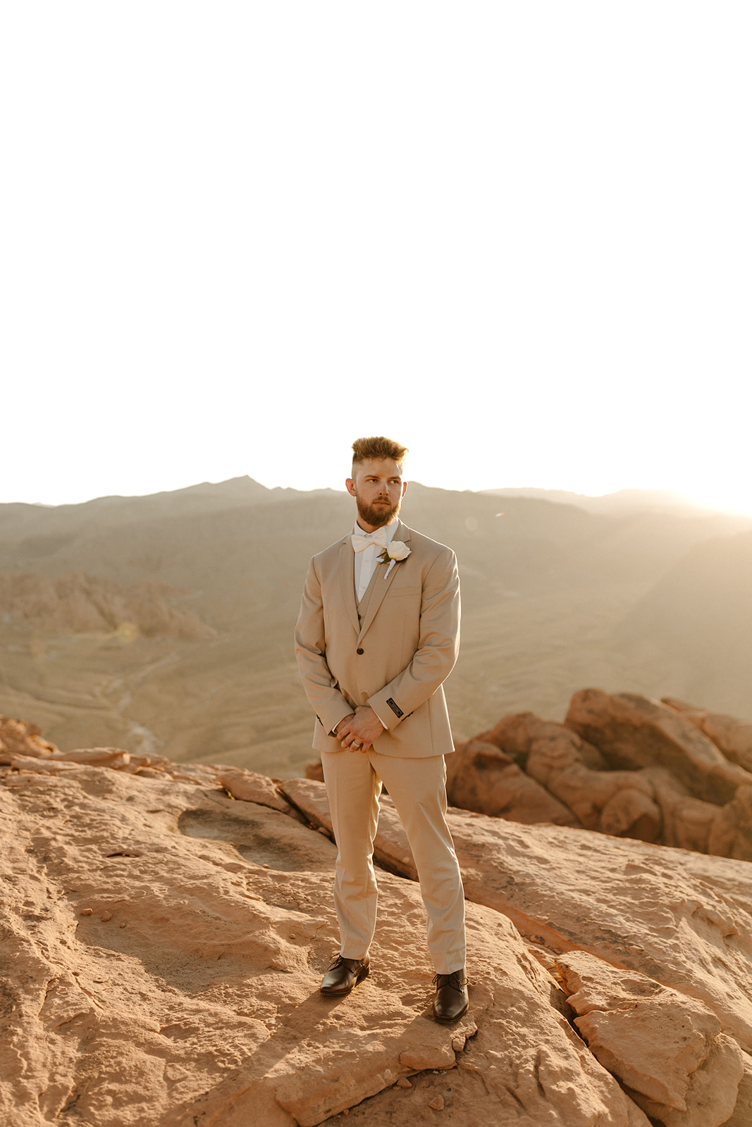 Golden hour desert portrait of groom wedding attire in a beige suit and white bow tie, standing solo on the rocky cliffside.