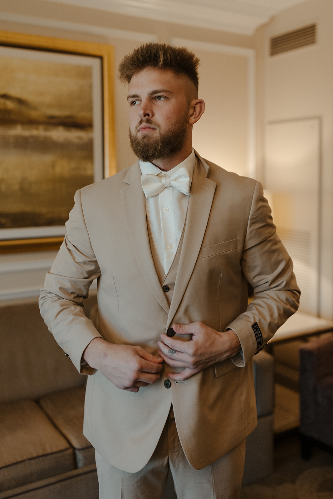 Groom wedding attire in a beige three-piece suit and ivory bow tie, buttoning up before the ceremony in a cozy hotel suite.
