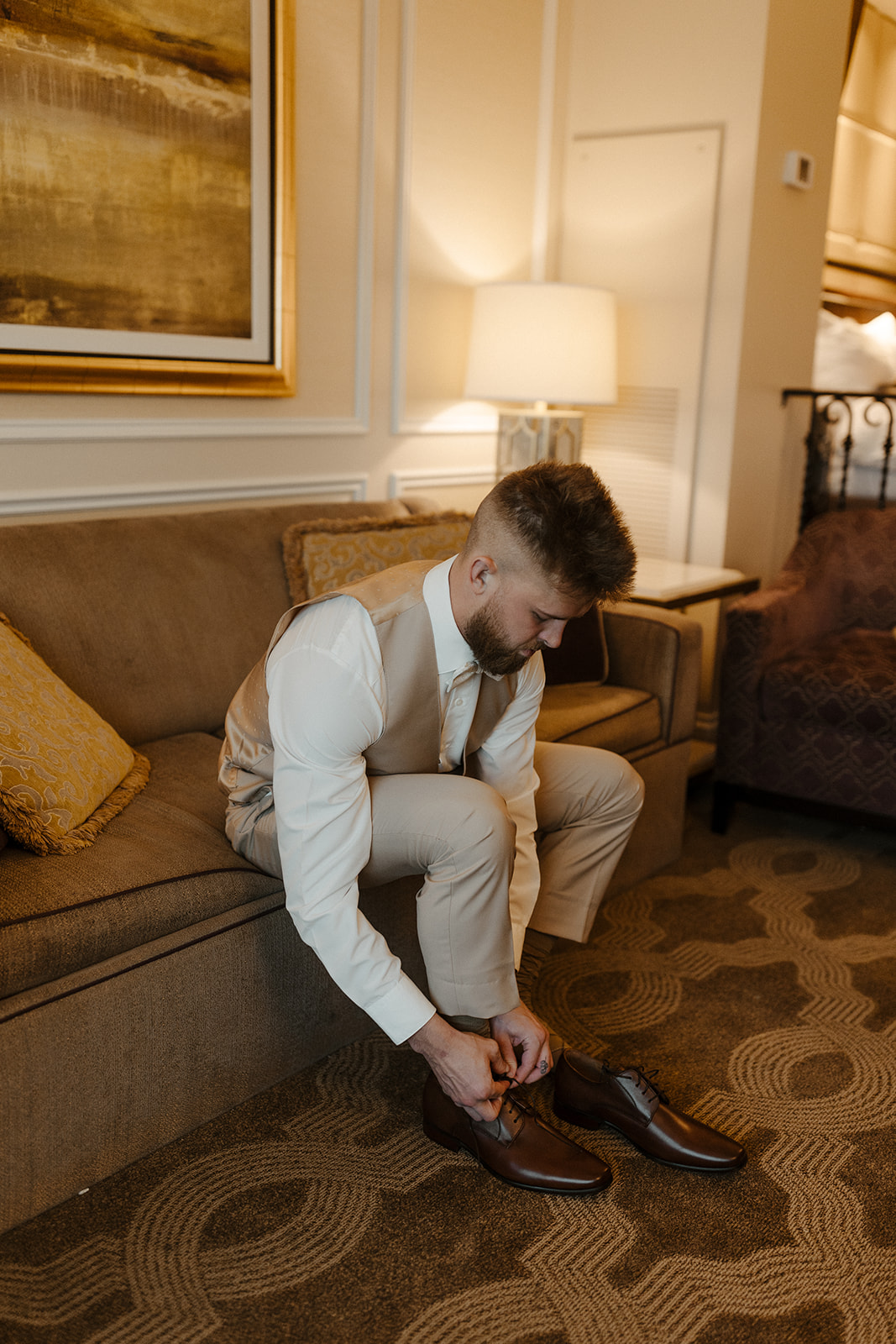 A groom in beige wedding attire laces up his brown leather shoes in a cozy hotel room before the ceremony—warm, timeless groom style.
